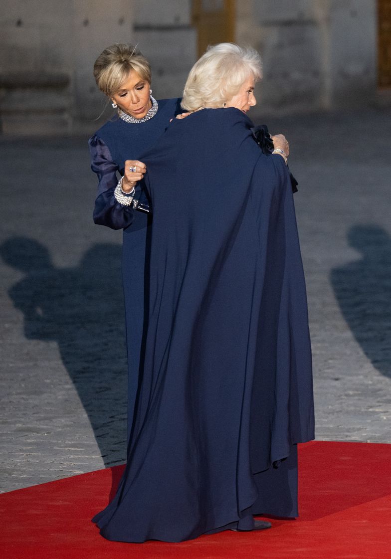 Pomocna Brigitte Macron poprawia pelerynę królowej Camilli. Panie postawiły na suknie wieczorowe od Diora