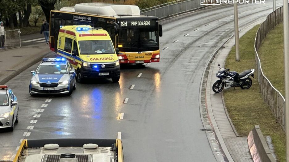 Na skrzyżowaniu zatrzymane są pojazdy: karetka z włączonymi sygnałami, radiowóz oraz autobus linii 194. Obok stoi motocykl.