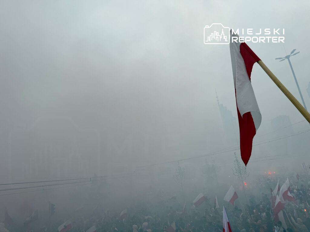 Tłum ludzi z flagami w chmurze dymu, w tle nowoczesne budynki.