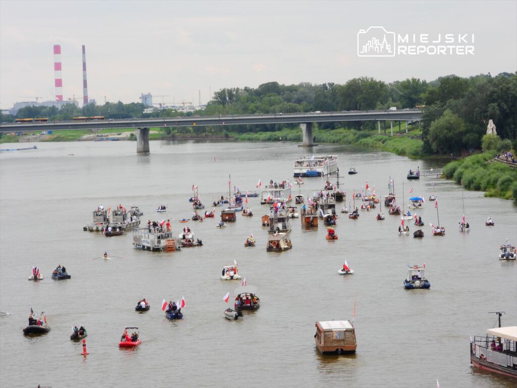 Na rzece pływa wiele łodzi, w tym motorówki i tradycyjne barki, z flagami. W tle widoczny most oraz przemysłowe budynki.