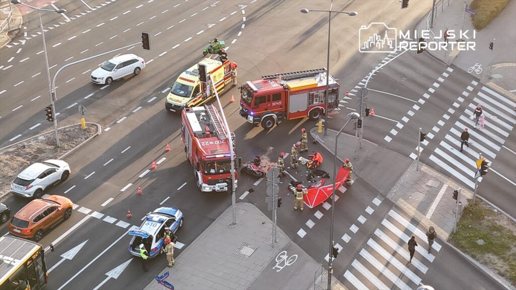 Na skrzyżowaniu ulic pracują strażacy i ratownicy medyczni, zabezpieczając miejsce wypadku motocyklowego.