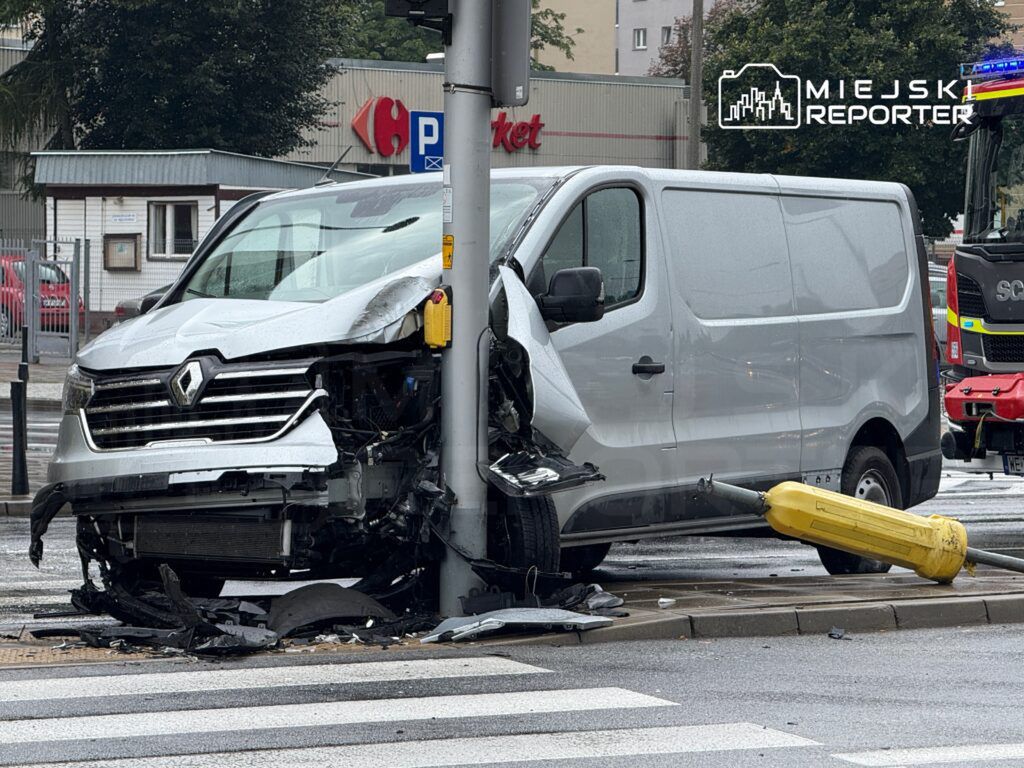 Uszkodzony biały furgon Renault uderzył w słup sygnalizacji świetlnej na deszczowej ulicy, obok sklepu.