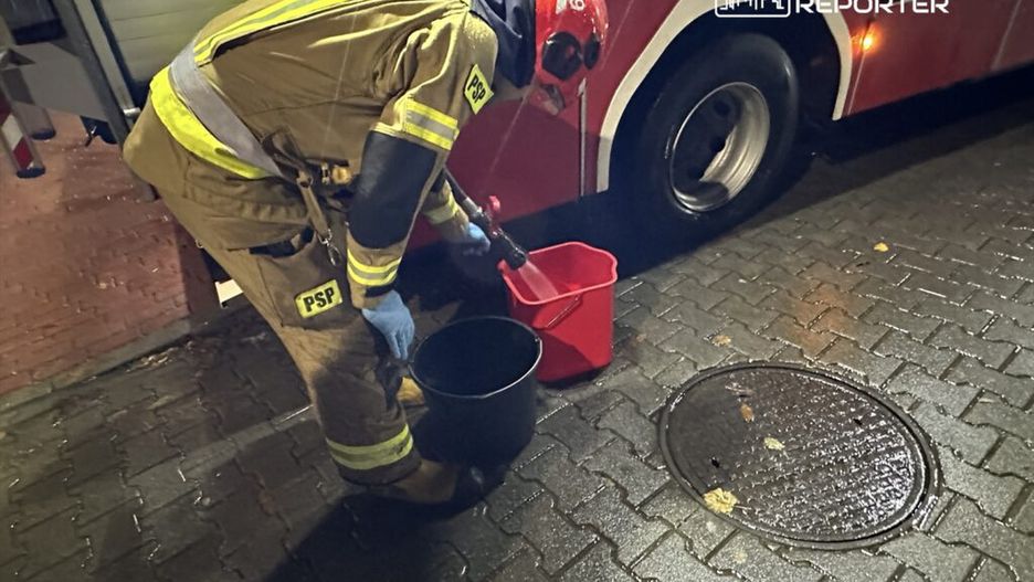 Strażak w mundurze z logo PSP nalewa wodę z czarnego wiadra do czerwonego pojemnika obok wozu strażackiego.