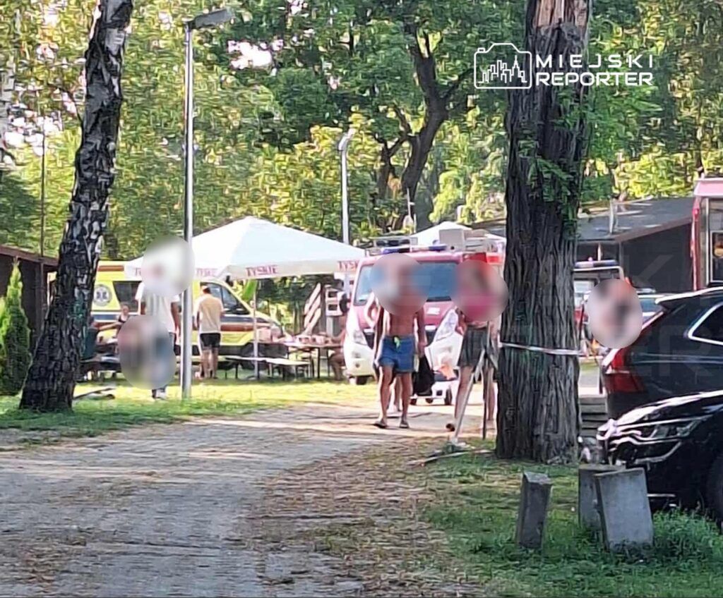 Grupa ludzi w letnich ubraniach przebywa w parku, obok namiotu z napisem "TYSIĄC". W tle widoczne karetki pogotowia i samochody.