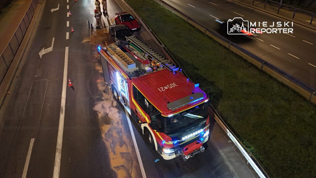 Wozidło strażackie z włączonymi sygnałami świetlnymi stoi na drodze, obok grupy strażaków oraz znaku ostrzegawczego.