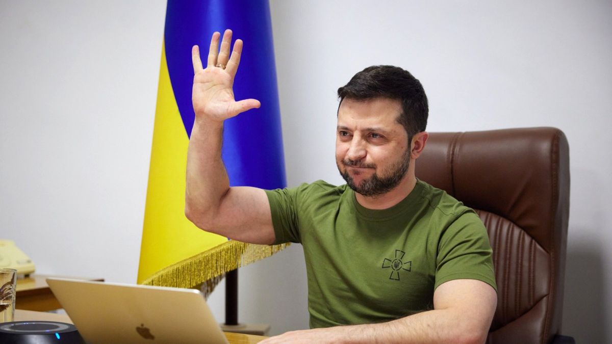 Na zdjęciu prezydent Ukrainy Wołodymyr Zełenski przed laptopem unoszący dłoń. Za nim flaga Ukrainy
