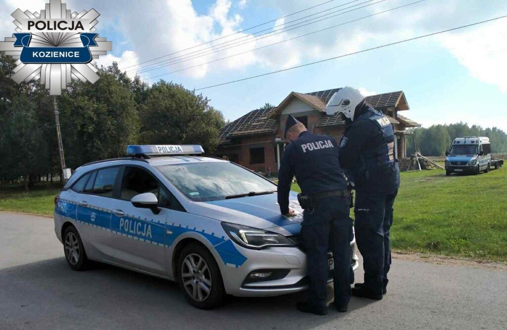 Dwaj policjanci w mundurach stoją obok radiowozu, rozmawiając w pobliżu budowy domu.
