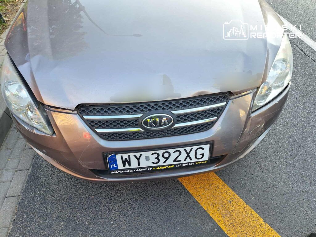 Kia z widoczną uszkodzoną karoserią zaparkowana na drodze, na tle asfaltu z wyraźnymi liniami.