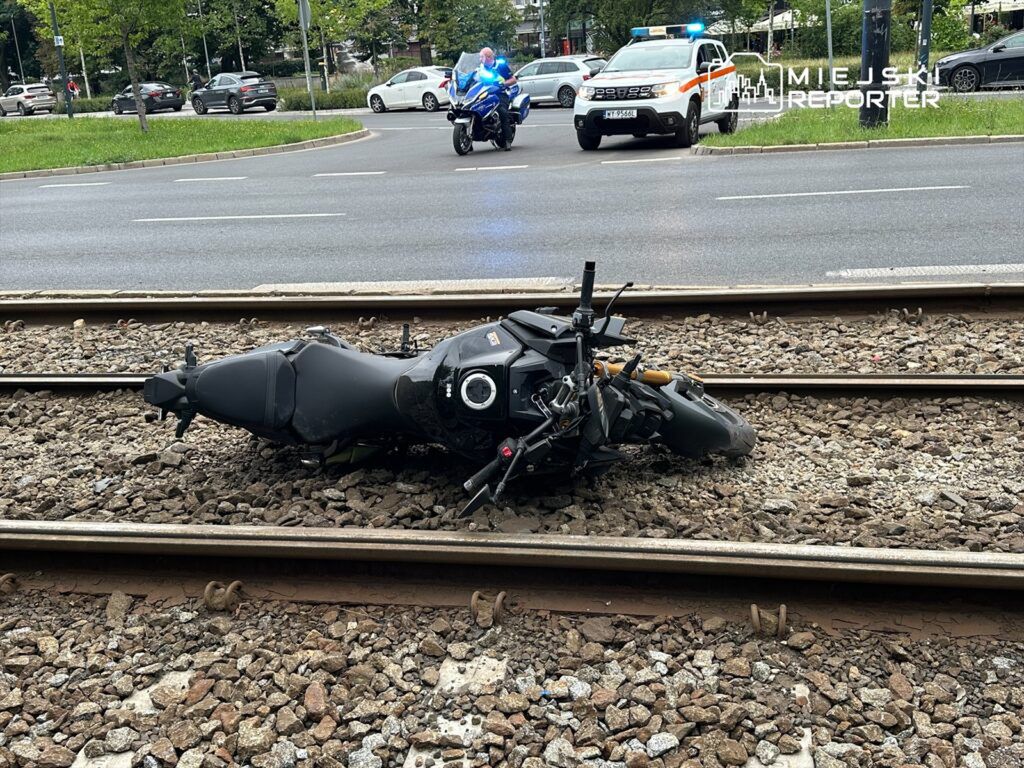 Motocykl leży na torach tramwajowych, a w tle widać radiowóz policyjny z włączonymi sygnałami świetlnymi.