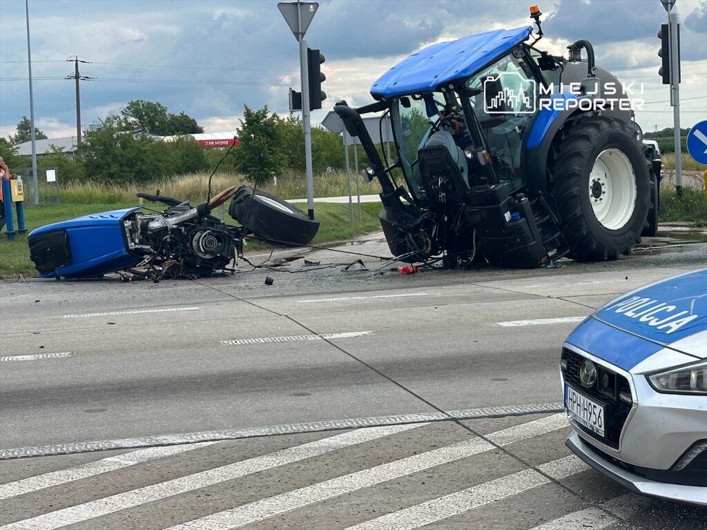 Traktor leży na boku na drodze, obok widać policyjny radiowóz oraz uszkodzone elementy maszyny.