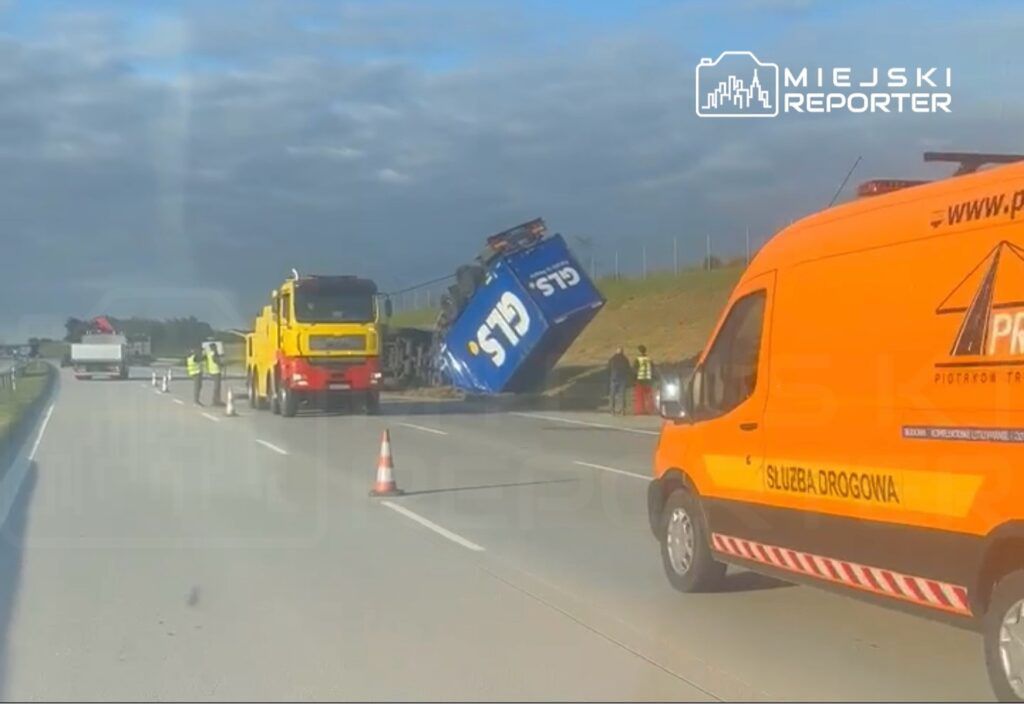 Czerwono-żółta ciężarówka z podnośnikiem stabilizuje przewróconą niebieską ciężarówkę na autostradzie.