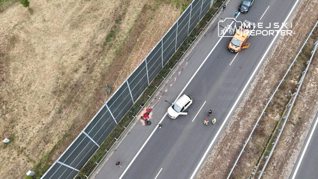 Wypadek drogowy z białym samochodem na poboczu, obok leży uszkodzony motocykl, a obok stoją ratownicy i świadkowie.