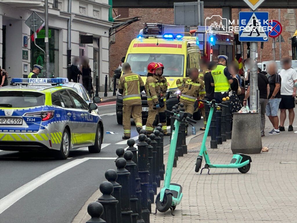 Grupa strażaków i policjantów interweniuje na ulicy, obok zaparkowanej karetki pogotowia. Na chodniku stoją elektryczne hulajnogi.
