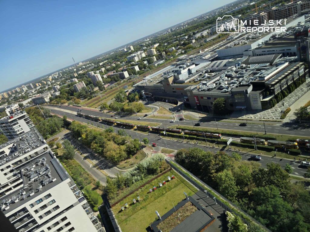 Widok z góry na ruchliwą ulicę z tramwajami, w tle nowoczesny kompleks handlowy i zabudowa mieszkalna.
