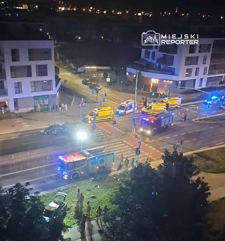 Na skrzyżowaniu nocą zablokowane pojazdy służb ratunkowych, w tym karetki i wóz strażacki, otoczone przez gapiów.