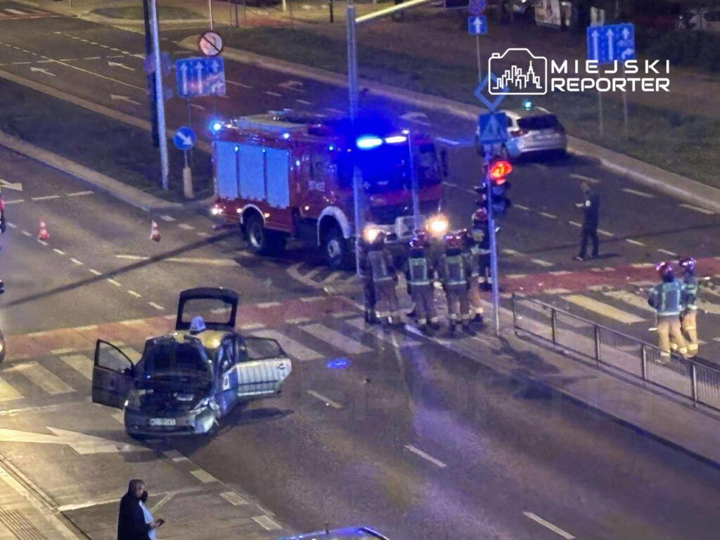 Strażacy w mundurach rozmawiają przy uszkodzonym samochodzie na skrzyżowaniu w nocy, obok wozu strażackiego.