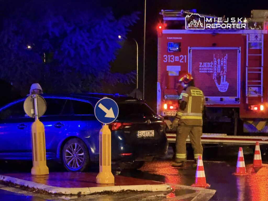 Strażak w mundurze obok wozu strażackiego, zabezpieczający miejsce zdarzenia w nocy, z oznaczeniami drogowymi w tle.