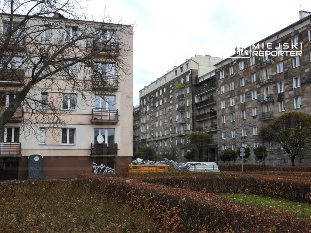 Nowoczesny budynek mieszkalny z balkonami oraz stary blok w tle, otoczone krzewami i drzewami w parku.