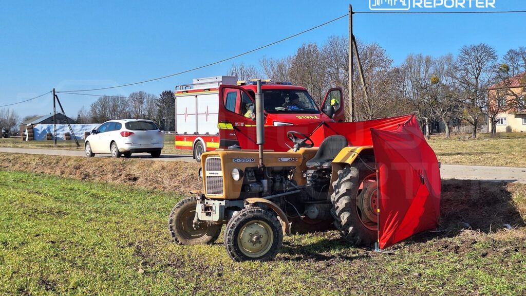 Traktor z czerwoną osłoną stoi na polu obok wozu strażackiego, a w tle widać wiejską drogę i domy.