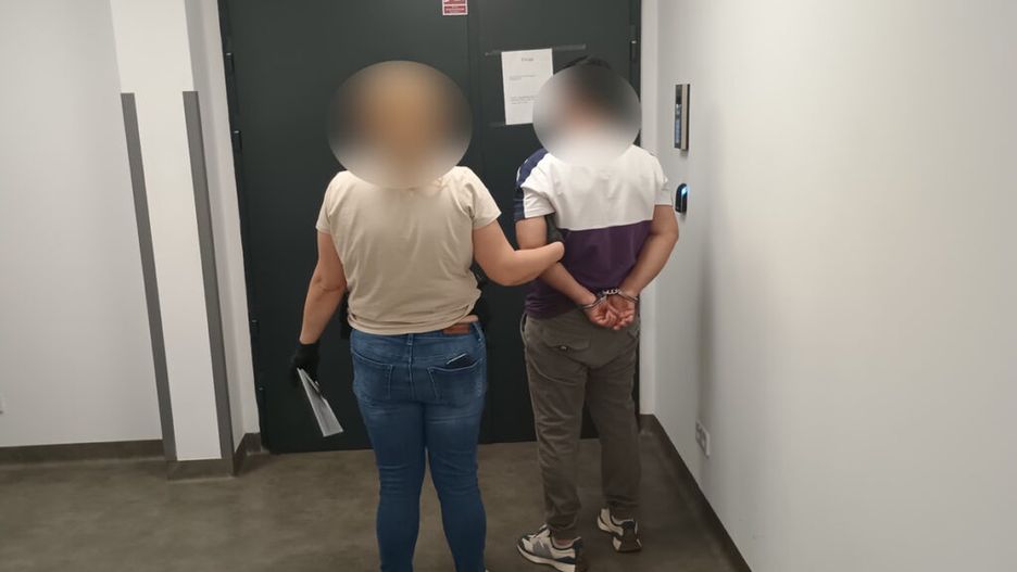 Kobieta w rękawiczkach prowadzi mężczyznę z rękami związanymi na plecach w korytarzu z szarymi ścianami.