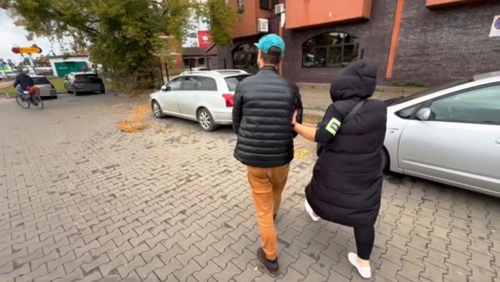 Mężczyzna w czarnej kurtce i kobieta w puchowej kurtce idą chodnikiem w pobliżu zaparkowanych samochodów.