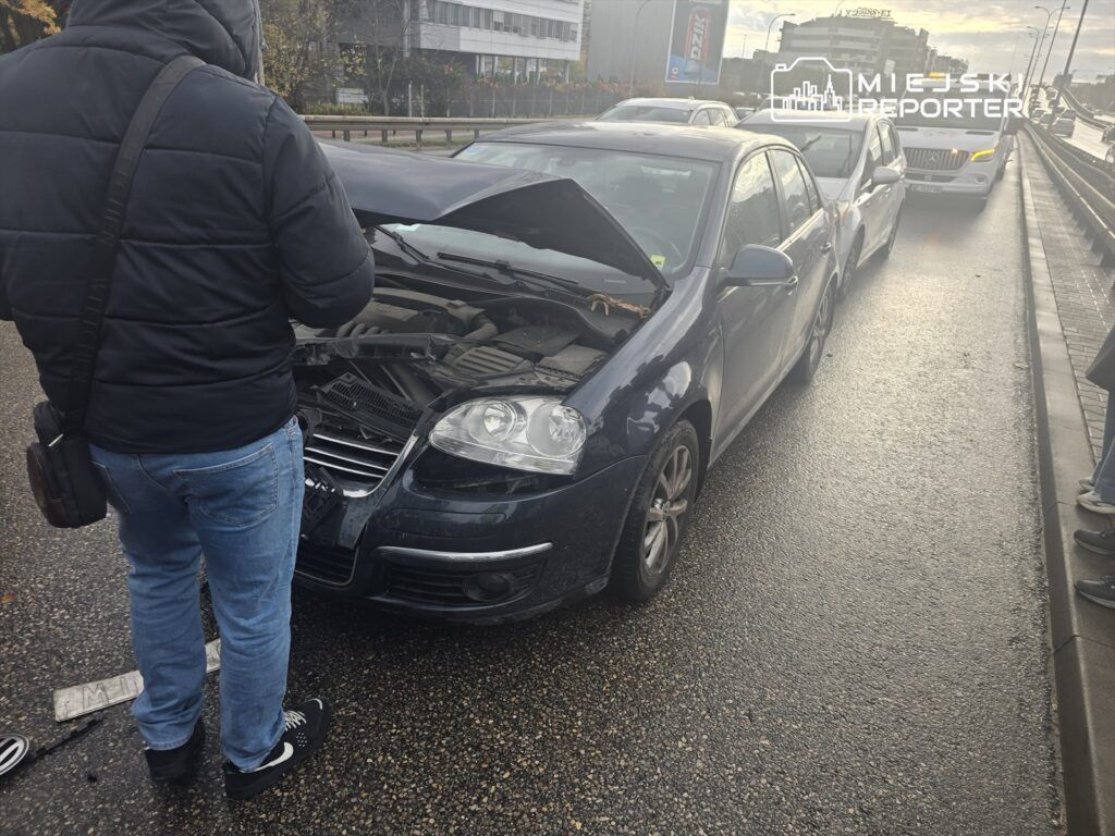 Mężczyzna w kurtce sprawdza silnik samochodu na ruchliwej ulicy, obok stoją inne pojazdy.