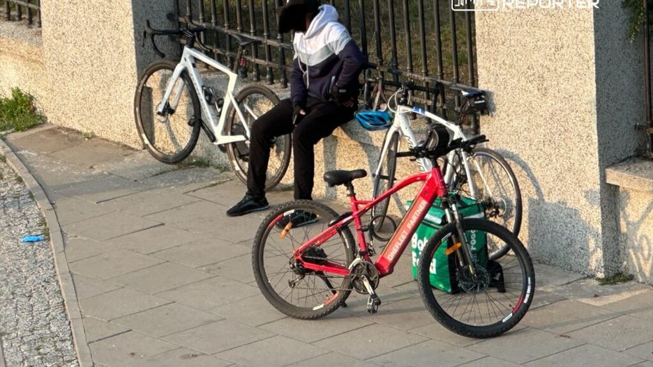 Mężczyzna w ciemnej odzieży siedzi obok dwóch rowerów na chodniku w miejskim otoczeniu.