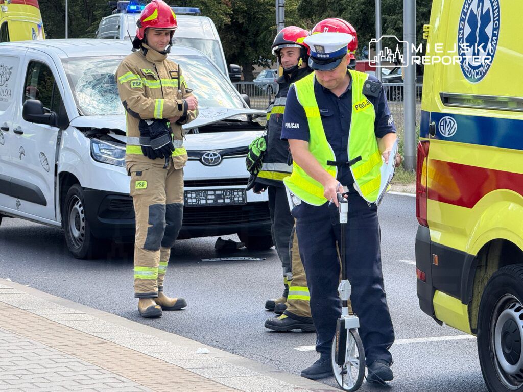 Strażak w mundurze z hełmem obserwuje miejsce wypadku, podczas gdy policjant mierzy odległość na jezdni. W tle uszkodzony samochód dostawczy.