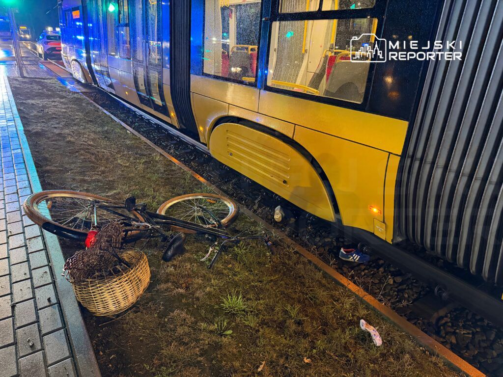 Przewrócony rower leży na trawie obok torów tramwajowych, w tle widać tramwaj oraz samochody w nocy.