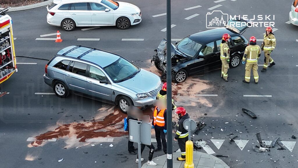 W miejscu wypadku drogowego, strażacy rozmawiają z osobami poszkodowanymi, a obok leżą resztki pojazdów.