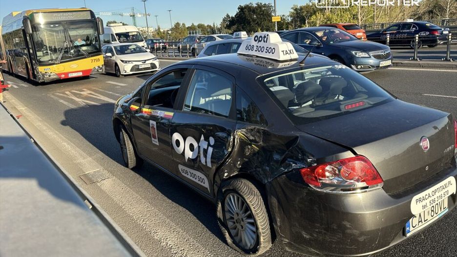 Czarny taksówka z widocznymi uszkodzeniami stoi na ruchliwej ulicy obok autobusu komunikacji miejskiej.