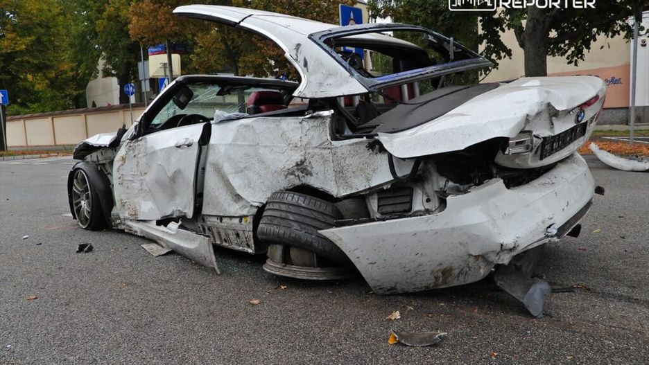 Zniszczony samochód osobowy leży na jezdni, z uszkodzeniami nadwozia i wygiętymi drzwiami, w otoczeniu drzew i znaków drogowych.