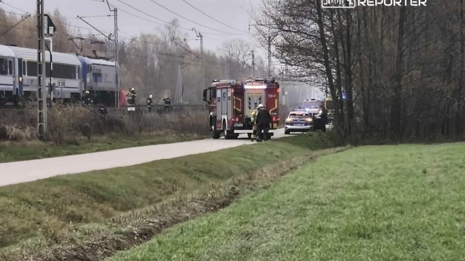 Strażacy i pojazdy ratunkowe pracują przy torach kolejowych w lesistym terenie, obok jeżdżącego pociągu.