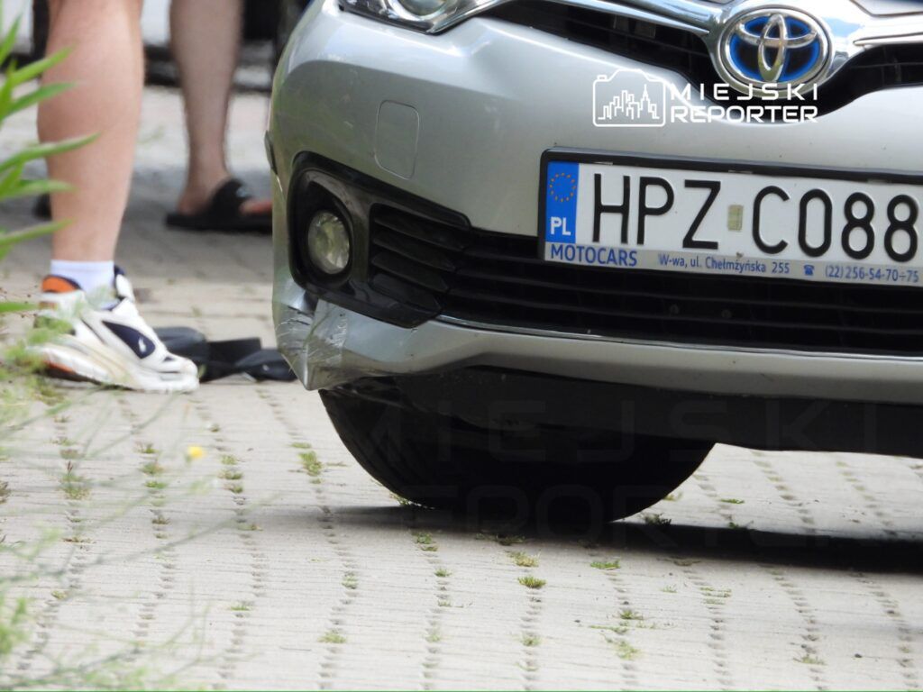 Samochód osobowy marki Toyota z uszkodzoną przednią częścią zaparkowany na chodniku w otoczeniu zieleni.