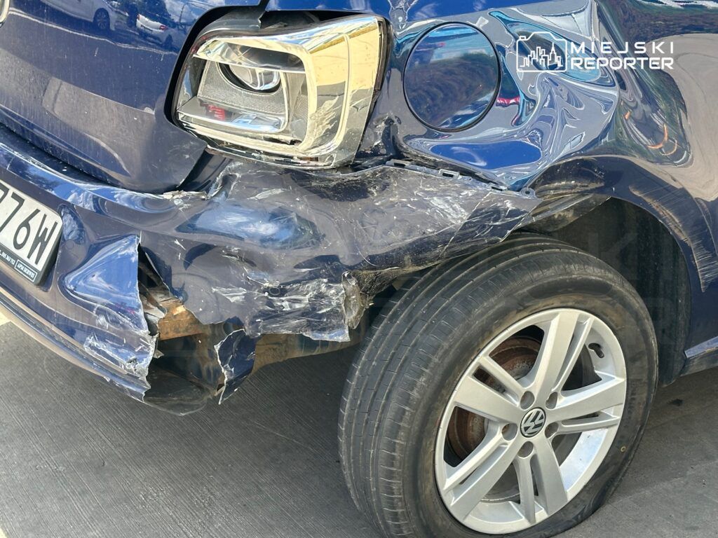 Uszkodzony niebieski samochód osobowy z wgniecioną przednią częścią i uszkodzoną oponą na parkingu.