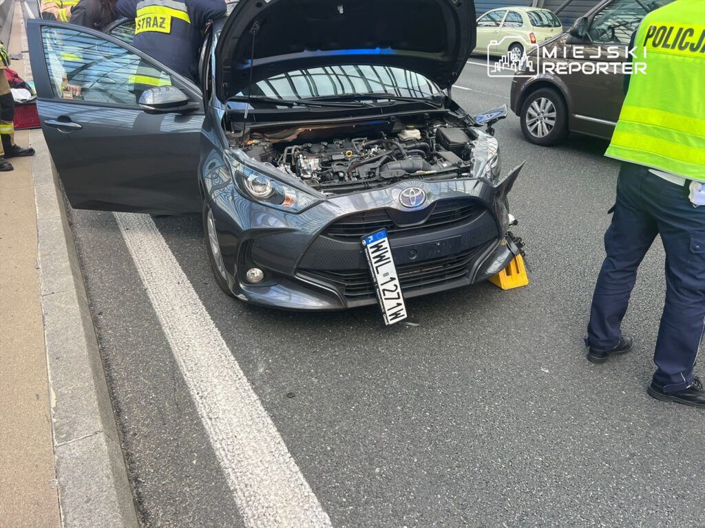 Toyota z otwartą maską stoi na drodze, obok niej pracują strażacy i policjant w kamizelce odblaskowej.