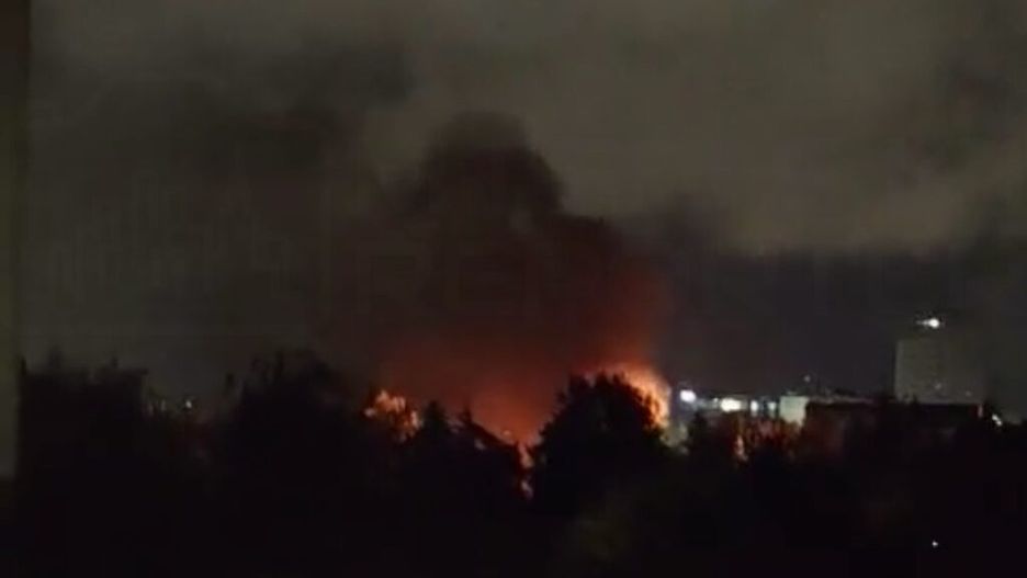 Płomienie i dym unoszące się nad drzewami w nocnym krajobrazie miejskim.