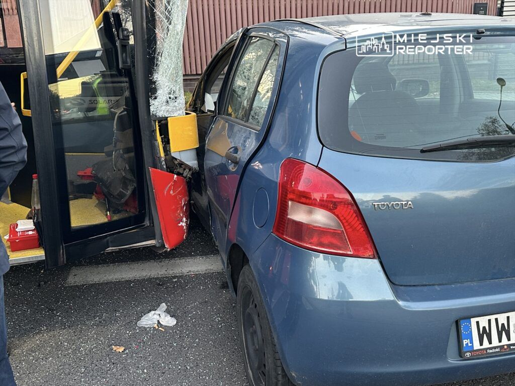 Zniszczony niebieski samochód osobowy uderzony w drzwi przez przystanek autobusowy, na tle miejskiej ulicy.
