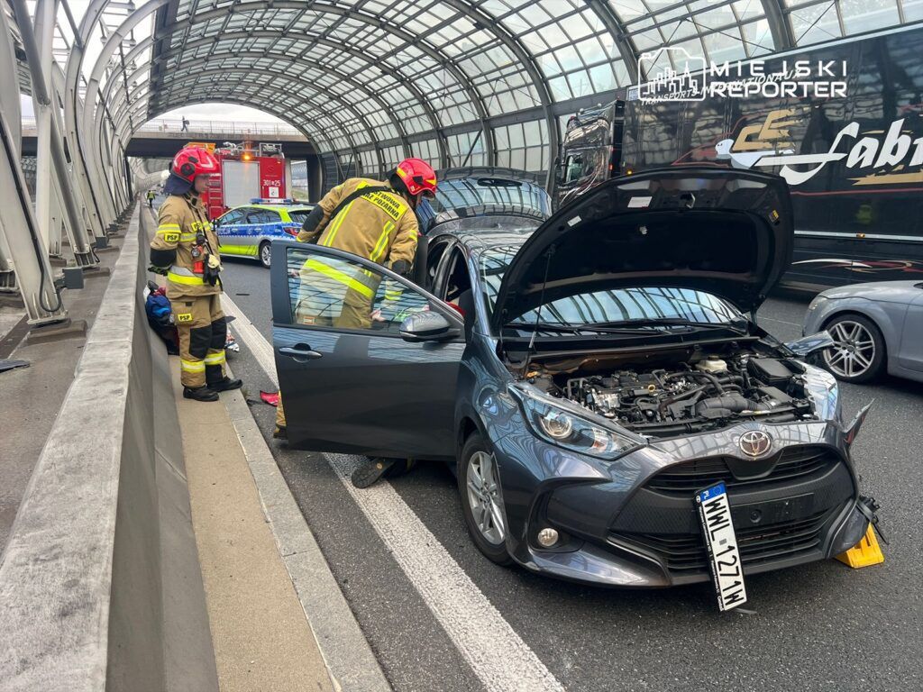 Dwaj strażacy w mundurach sprawdzają silnik samochodu osobowego na ruchliwej drodze, obok stoi wozem strażackim.