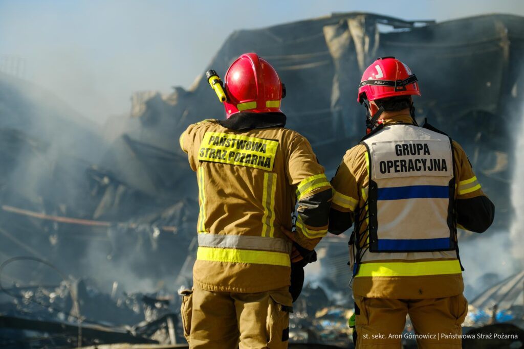 Dwaj strażacy w mundurach obserwują zgliszcza pożaru, jeden z nich wskazuje na miejsce zdarzenia.