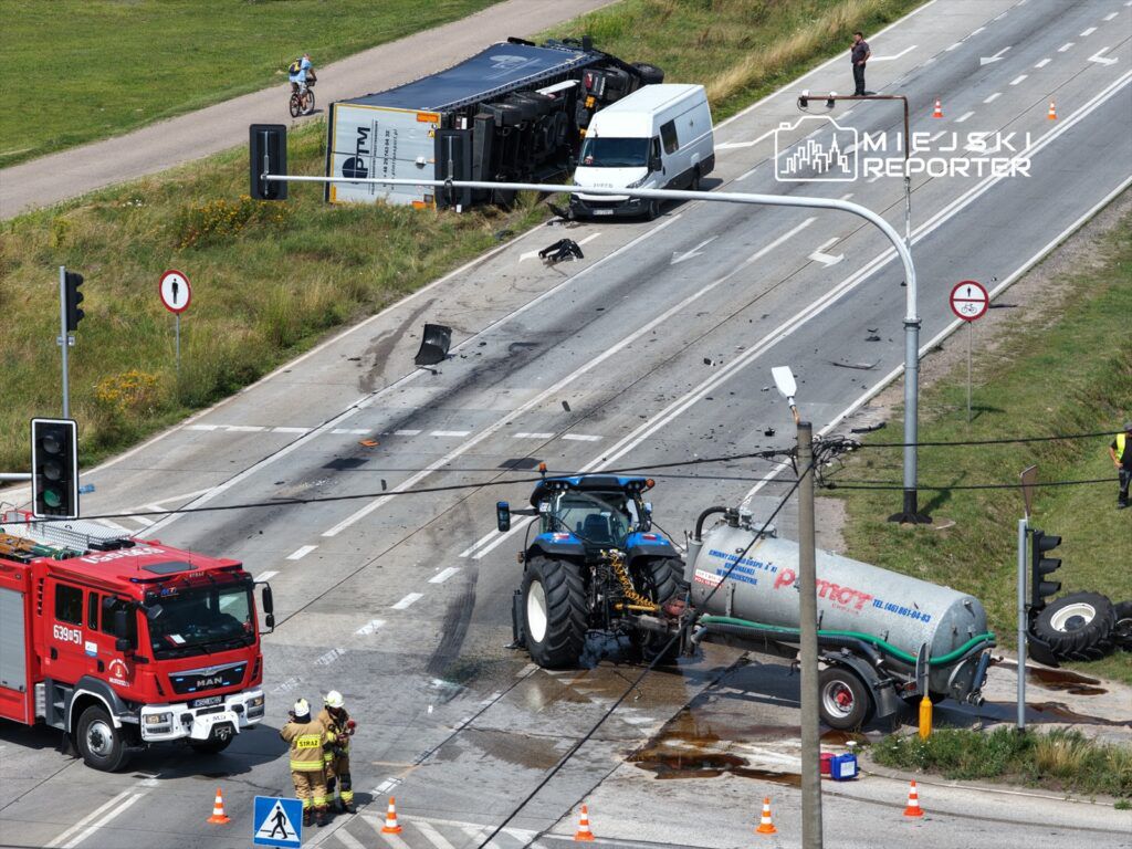 W wyniku wypadku na drodze leży przewrócony ciężarówka, a obok stoi traktor z przyczepą. W pobliżu interweniują strażacy.