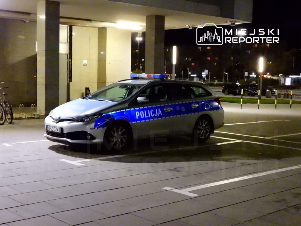 Policyjny samochód zaparkowany na nocnym parkingu przed budynkiem, z włączonymi światłami.