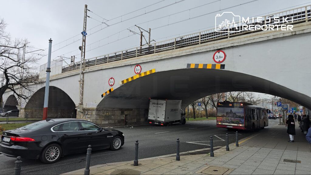 Samochód osobowy i ciężarówka przejeżdżają pod mostem kolejowym z widocznymi znakami wysokościowymi.