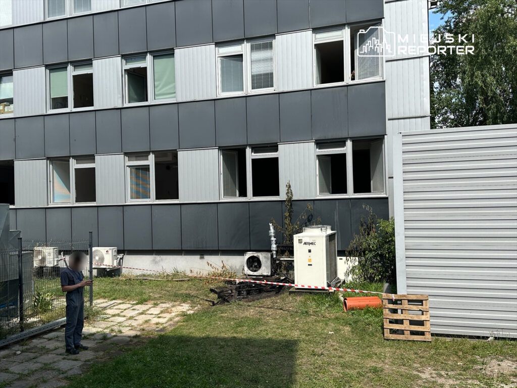 Mężczyzna w roboczym stroju stoi obok nowoczesnego budynku z otwartymi oknami, w tle widoczne uszkodzenia.