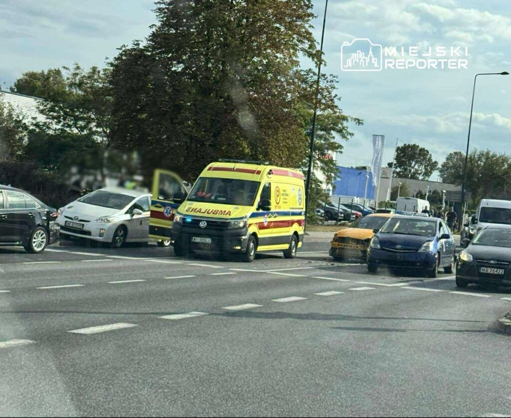 Karetka pogotowia z włączonymi sygnałami świetlnymi stoi w zatorze drogowym obok uszkodzonego samochodu.