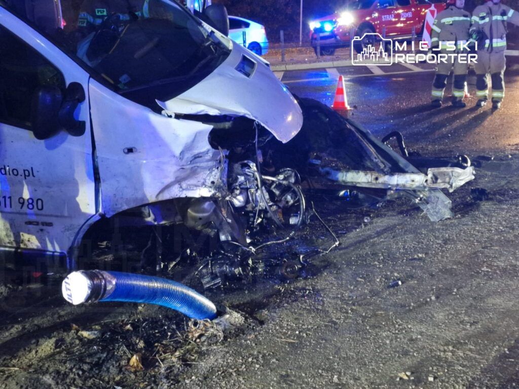 Uszkodzony pojazd dostawczy z wgniecioną przednią częścią stoi na drodze, obok interweniują strażacy.