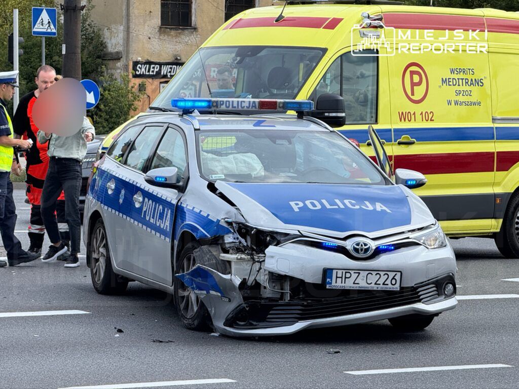 Policyjny radiowóz z uszkodzoną przednią częścią stoi na jezdni, obok funkcjonariusze rozmawiają z osobą poszkodowaną. W tle widać ambulans.