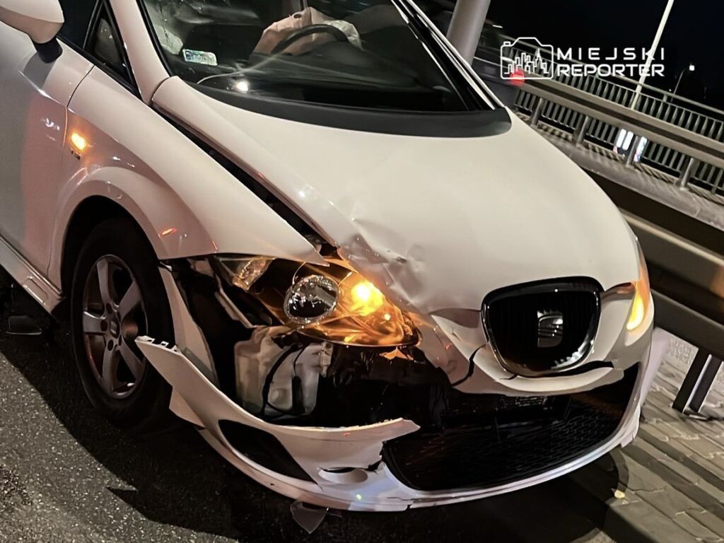 Uszkodzony biały samochód osobowy zaparkowany na drodze, z włączonymi światłami awaryjnymi.