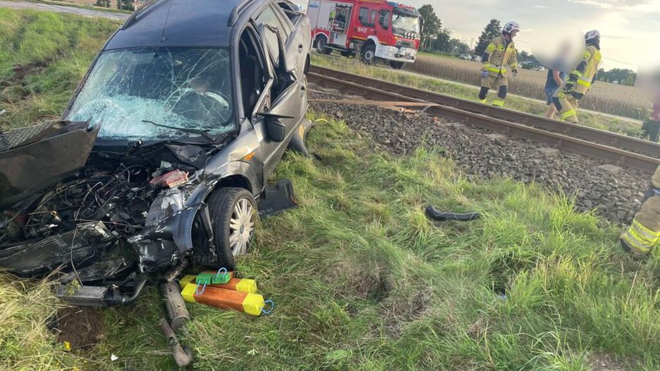 Uszkodzony samochód leży na poboczu drogi obok torów kolejowych, a strażacy udzielają pomocy osobie poszkodowanej.