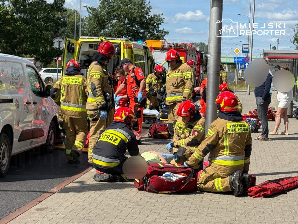 Zespół ratowników medycznych i strażaków udziela pomocy poszkodowanemu na chodniku obok zaparkowanej karetki.